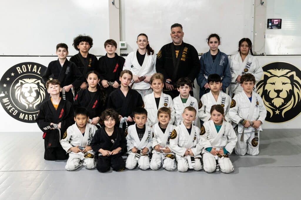 young students and instructors posing for photo in front of logo banner for kids jiu jitsu class at Royal Mixed Martial Arts
